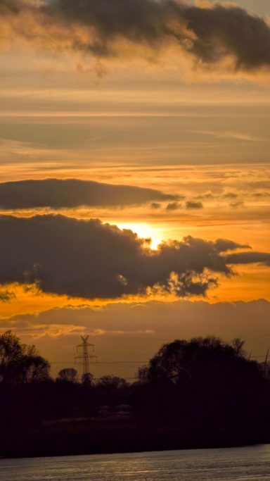 Sonnenuntergang hinter Wolken, orange und gelbe Töne am Himmel über Silhouette von Bäumen.