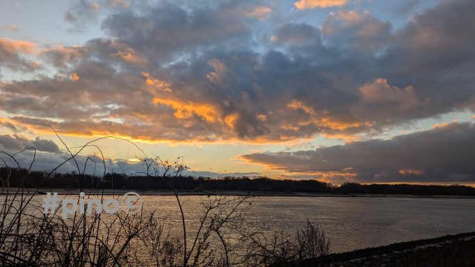 Sonnenuntergang über einem Fluss mit bewölktem Himmel und Silhouette von Bäumen.