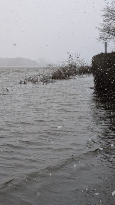 Überflutetes Ufer mit trübem Wasser und leichten Schneeflocken im Fall.