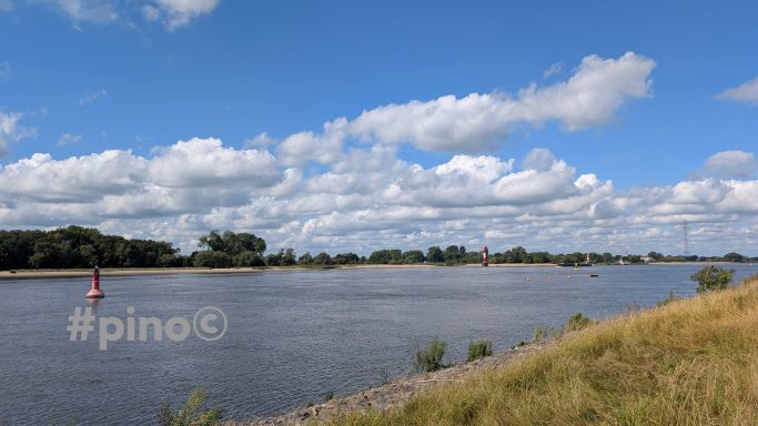 Blauer Himmel mit Wolken über einem ruhigen Fluss, gesäumt von Ufervegetation.