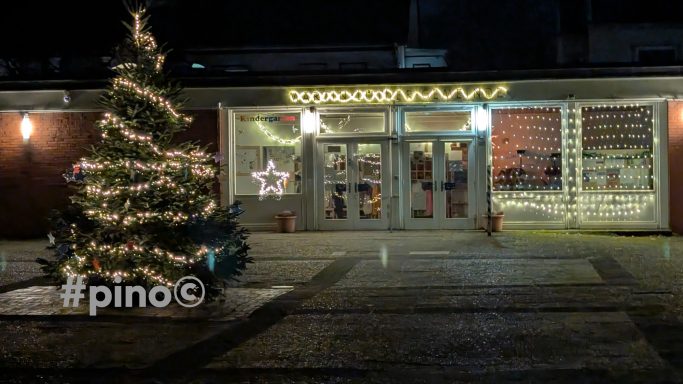 Weihnachtlich dekorierte Fassade mit einem beleuchteten Baum und festlichen Lichtern.