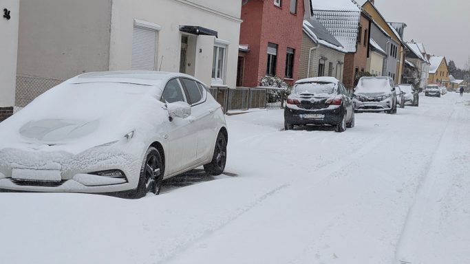 Zwei Autos auf einer verschneiten Straße, umgeben von Häusern und Schnee.