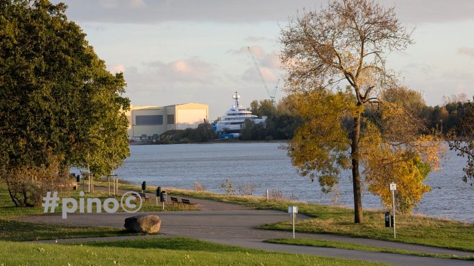 Herbstliche Parklandschaft am Wasser mit Bäumen und Spaziergängern, im Hintergrund ein Gebäude.
