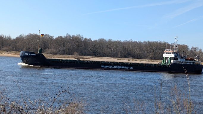 Schwarzes Frachtschiff auf einem Fluss mit Bäumen im Hintergrund.
