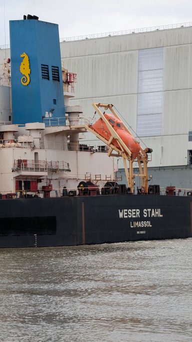 Containerschiff mit Kran und orangefarbener Ladeeinheit, angedockt an einem Hafen.