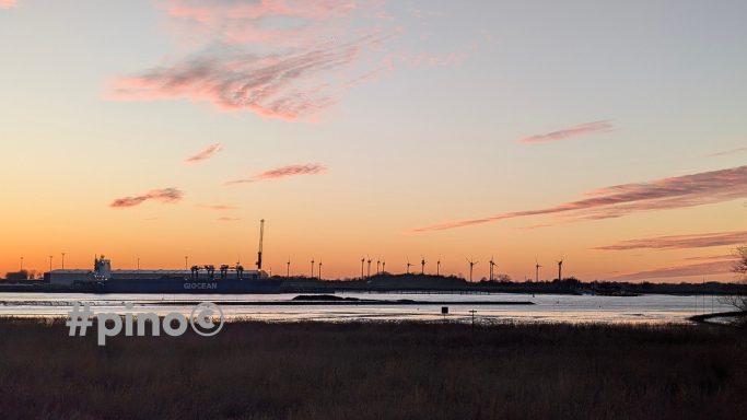 Sonnenuntergang über einem Gewässer mit Silhouetten von Windkraftanlagen im Hintergrund.