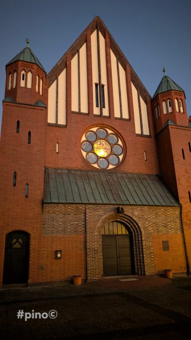 Fassade einer Kirche mit einem großen Fenster und drei Türmen bei Abendlicht.