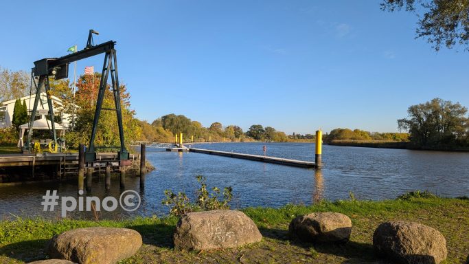 Ruhiger Fluss mit Ufer und Bäumen, Krananlage im Vordergrund, blauer Himmel.