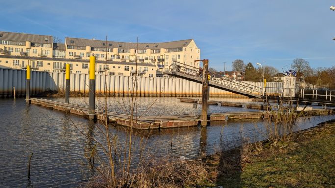 Ufer mit Gebäuden im Hintergrund, Steg und Wasserfläche bei Sonnenlicht.
