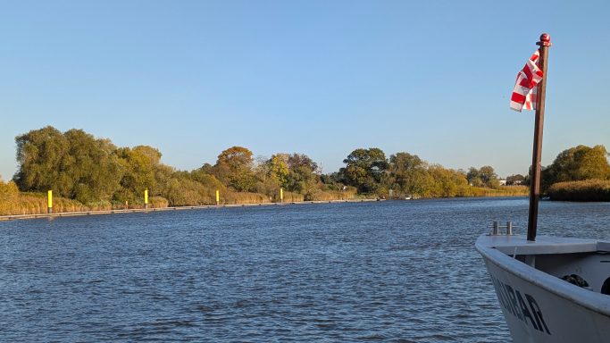 Blick auf einen Fluss mit Bäumen und einem Boot, das eine rote Flagge trägt.