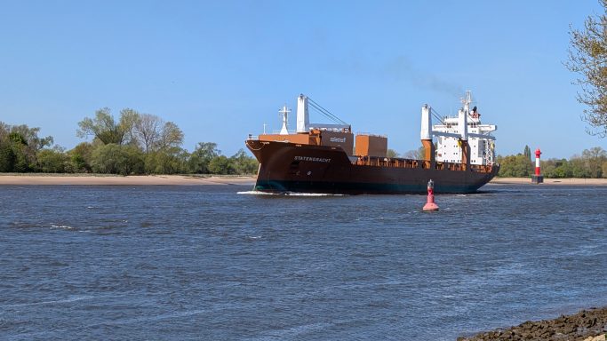 Ein Frachtschiff fährt auf einem Fluss mit grüner Ufervegetation.