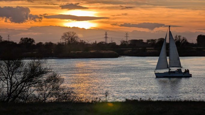 Segelboot auf ruhigem Wasser vor einem Sonnenuntergang mit orangefarbenem Himmel.