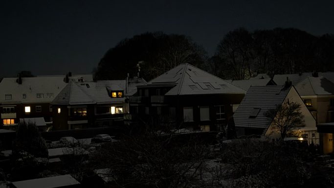 Schneebedeckte Dächer von Häusern bei Nacht mit wenigen beleuchteten Fenstern.