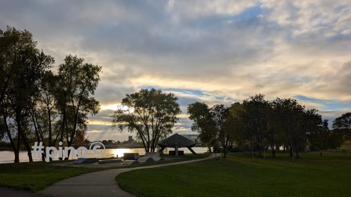 Parkanlage mit Wegen, Bäumen und einem Pavillon am Wasser bei Sonnenuntergang.