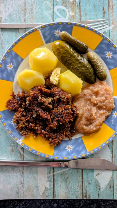 Ein Teller mit gekochten Kartoffeln, eingelegten Gurken, Apfelmus und Hackfleisch.