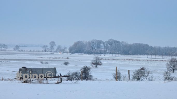Winterlandschaft mit verschneitem Feld und einem alten, verlassenen Gebäude im Hintergrund.