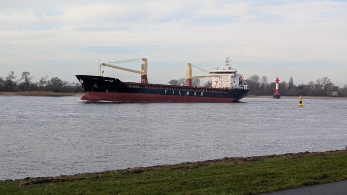 Ein Frachter fährt auf einem ruhigen Gewässer, umgeben von roten und gelben Hafenmarkierungen.