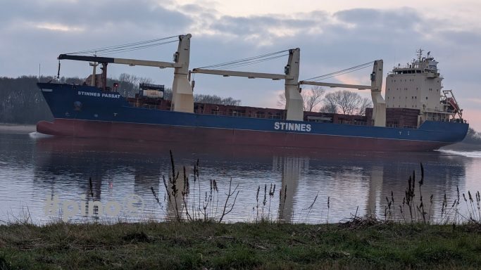 Ein Frachtschiff liegt an einem ruhigen Gewässer, umgeben von Bäumen und Wolken.