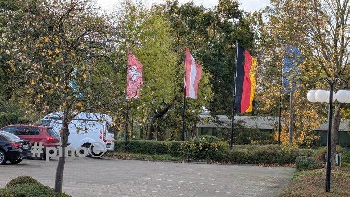 Drei Fahnen wehen vor einem Parkplatz mit Bäumen und Autos.