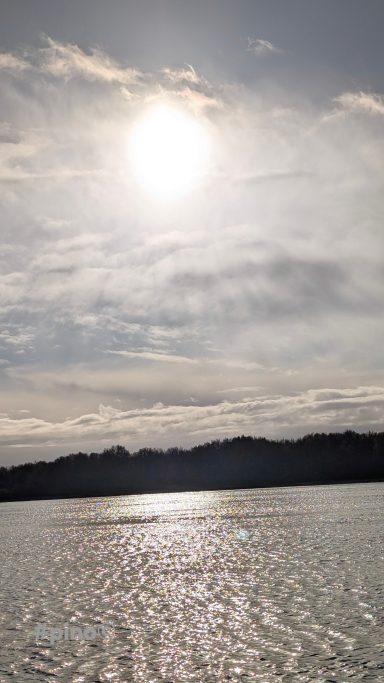 Helle Sonne über einem ruhigen Gewässer mit sanften Wellen und bewölktem Himmel.