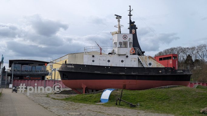 Ein schwarzes und gelbes Schlepperboot steht an Land, umgeben von Bäumen und Wolken.