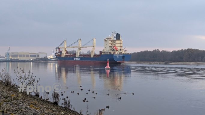 Frachtship fährt auf ruhigem Wasser, umgeben von Ufervegetation und einer sanften Landschaft.