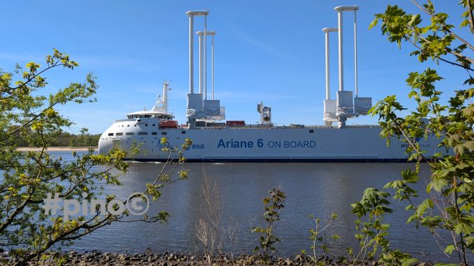 Ein Schiff mit zwei großen Kränen auf ruhigem Wasser, umgeben von grüner Vegetation.