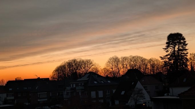 Sonnenuntergang über einem Stadtbild mit silhouettierten Bäumen und Wolken.