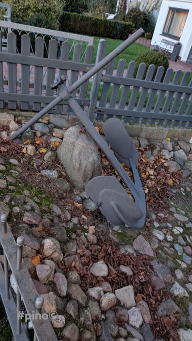 Ein Anker liegt zwischen Steinen und Laub hinter einem Holzzaun.