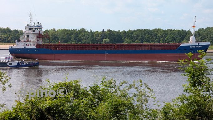 Ein Frachtschiff liegt am Ufer eines Flusses, umgeben von Bäumen und klarem Himmel.