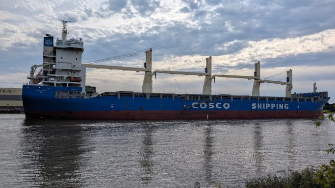 Großes Containerschiff mit COSCO-Logo fährt auf einem ruhigen Wasserweg.