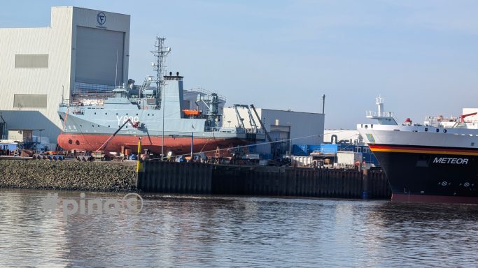 Schiffe im Hafen mit Lagerhäusern im Hintergrund, ruhiges Wasser.