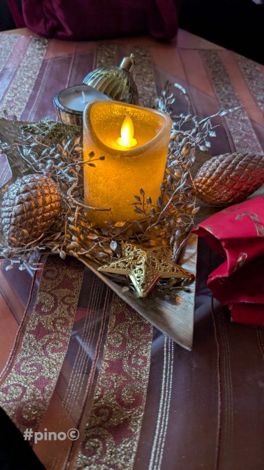 Dekorative Weihnachtsanordnung mit Kerze und goldenen Ornamenten auf rotem Tisch.