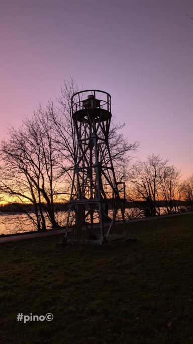 Blick auf einen Aussichtsturm am Wasser bei Sonnenuntergang, umgeben von Bäumen.